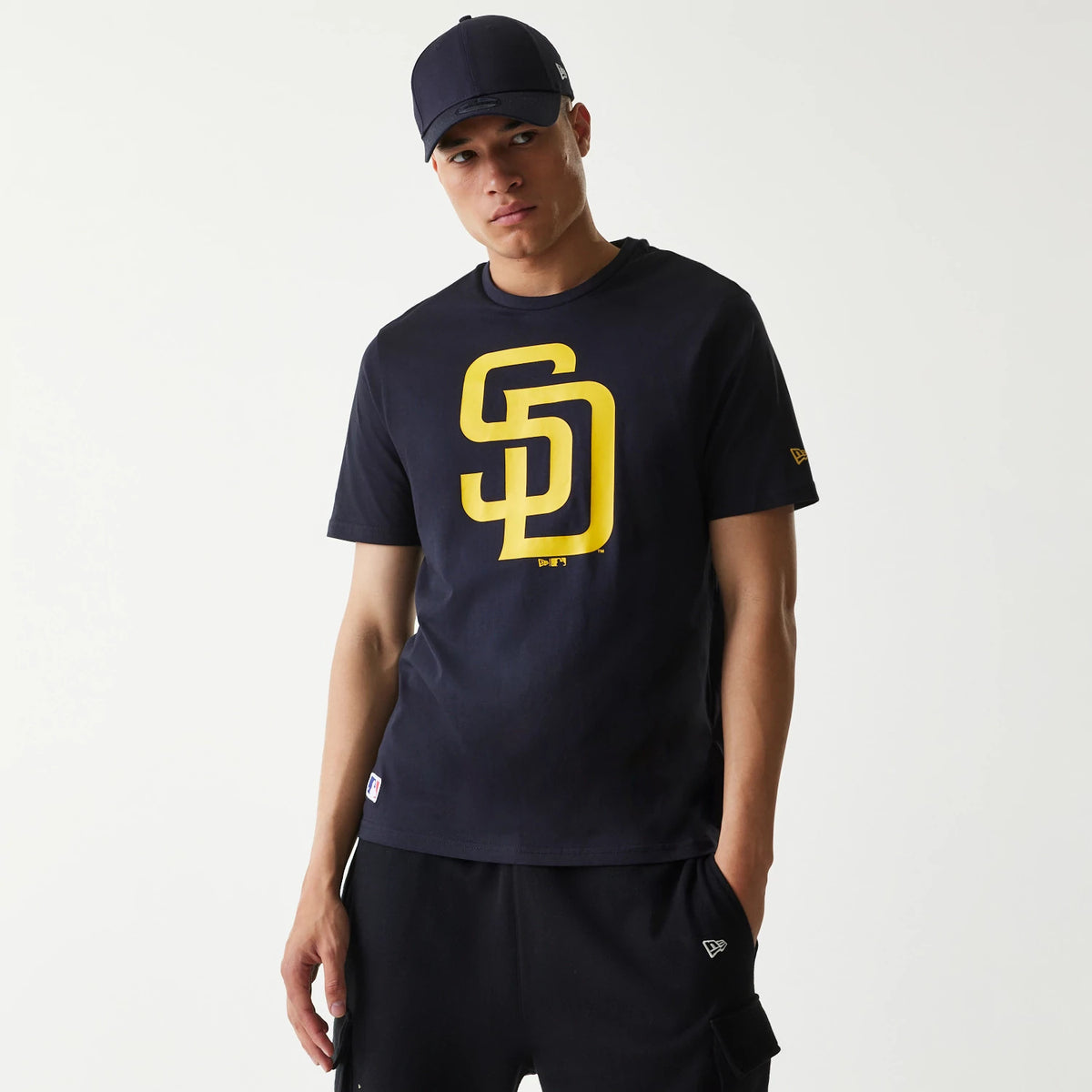 New Era San Diego Padres MLB Regular Navy T-Shirt, on-model view showing the navy shirt with a large yellow SD logo on the front, a small raised New Era logo on the left sleeve, and a black cap worn by the model.