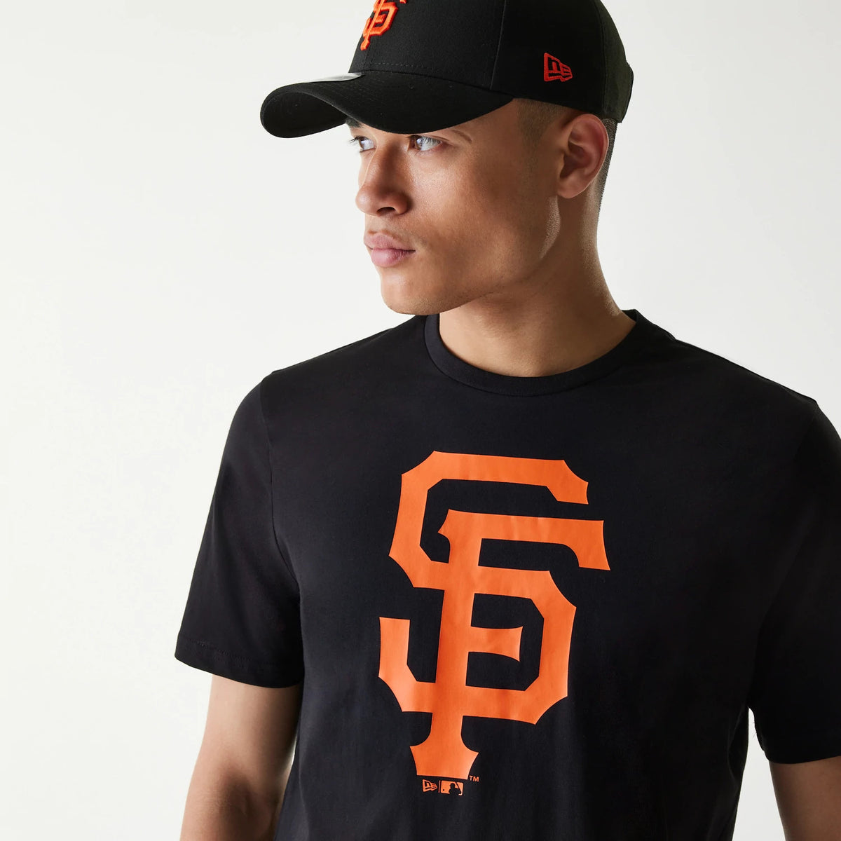 New Era San Francisco Giants MLB Regular Black T-Shirt, on-model view showing a large orange SF logo on the chest, worn with a black cap featuring a raised orange SF on the front and a debossed New Era logo on the side.