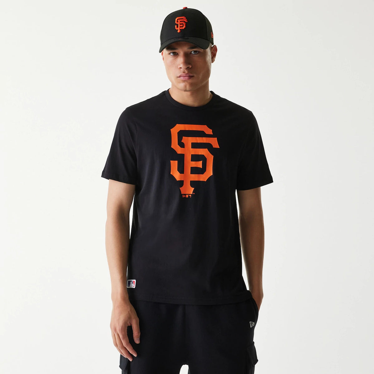 New Era San Francisco Giants MLB Regular Black T-Shirt, on-model view showing a large orange SF logo printed on the front centre of the black t-shirt, worn with a black cap featuring an embroidered orange SF.