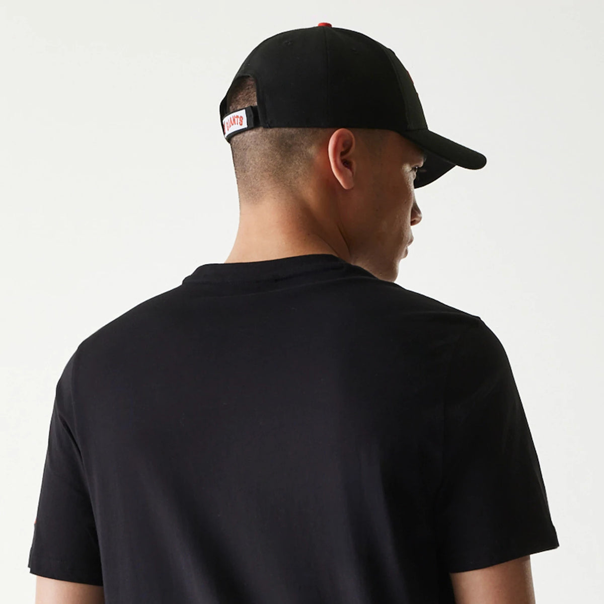 New Era San Francisco Giants MLB Regular Black T-Shirt, on-model view showing the black shirt from the back, the figure wearing a black cap with an adjustable strap, and the shirt's plain design highlighted from the rear.