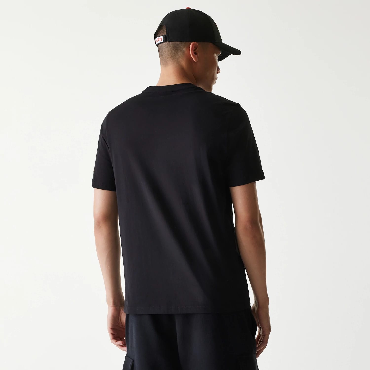 New Era San Francisco Giants MLB Regular Black T-Shirt, rear view showing the plain black back with short sleeves and a crew neckline, worn by a person with a black cap featuring a white embroidered logo above the adjustment strap.