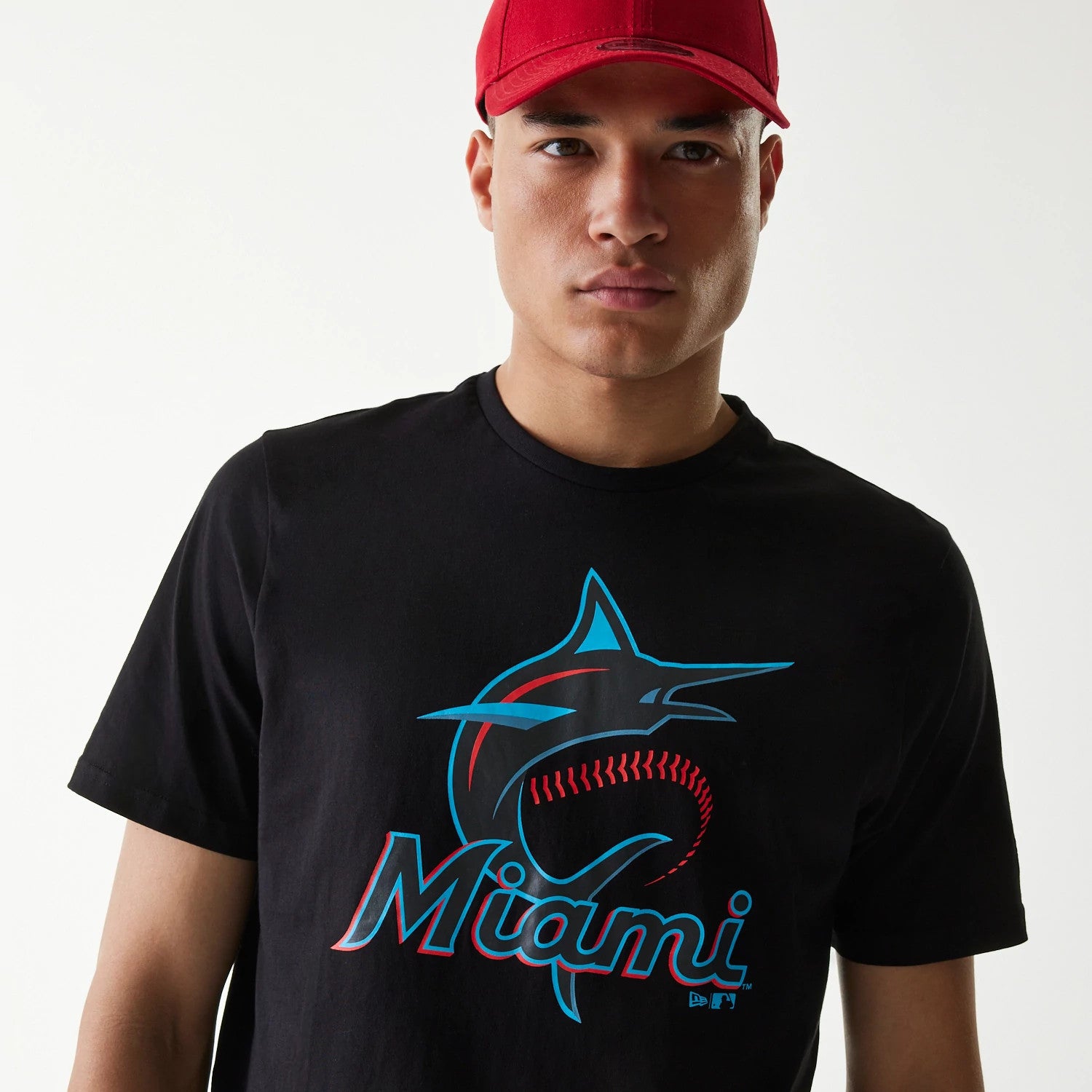 New Era Miami Marlins MLB Regular Black T-Shirt, front view showing a dark T-shirt with the Miami Marlins logo in red and blue across the chest, worn by a person also wearing a plain red cap.