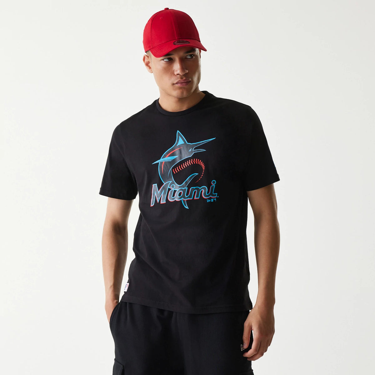 New Era Miami Marlins MLB Regular Black T-Shirt, on-model view showing the black shirt with a large marlin and baseball graphic on the chest in turquoise and red, worn by a person in a red cap with a debossed New Era logo on the front.