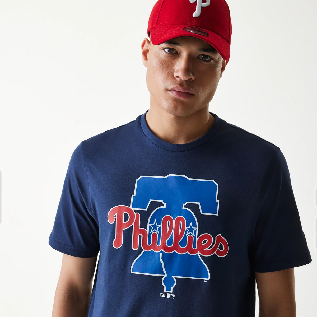 New Era Philadelphia Phillies MLB Regular Dark Blue T-Shirt, on-model showing a large red Phillies text logo with a blue Liberty Bell graphic on the front, paired with a red cap featuring a raised white P logo at the centre front.