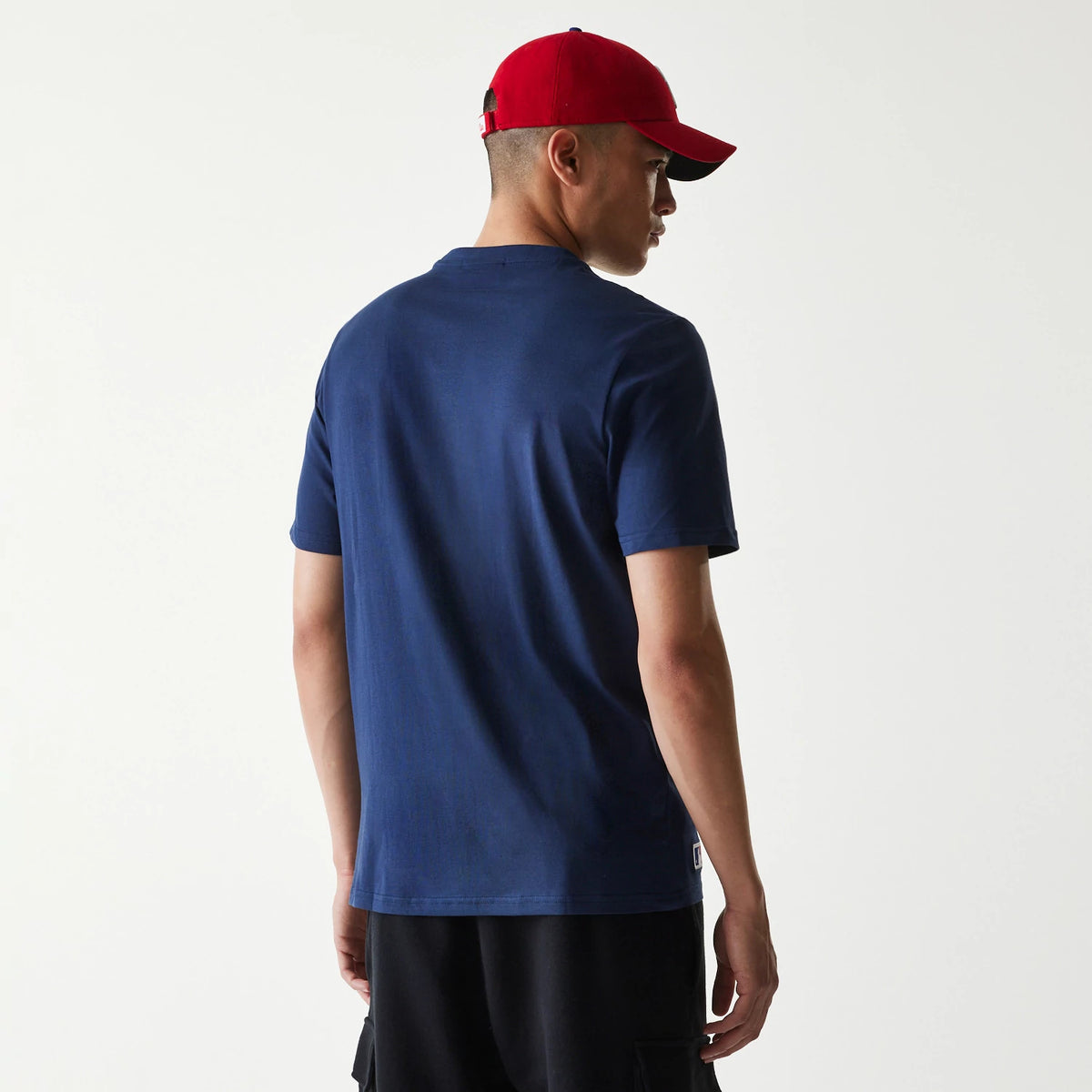 New Era Philadelphia Phillies MLB Regular Dark Blue T-Shirt, rear view showing the plain dark blue back with short sleeves worn by a model, a small raised logo tag at the lower hem on the left side, styled with a red cap.
