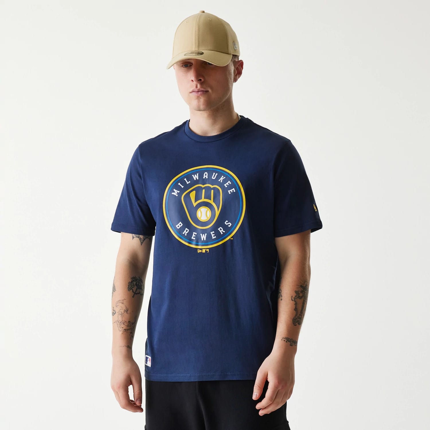 New Era Milwaukee Brewers MLB Regular Dark Blue T-Shirt, on-model view showing the circular Brewers logo centred on the chest in yellow and white, with a beige cap worn above and small logo detail on the sleeve.