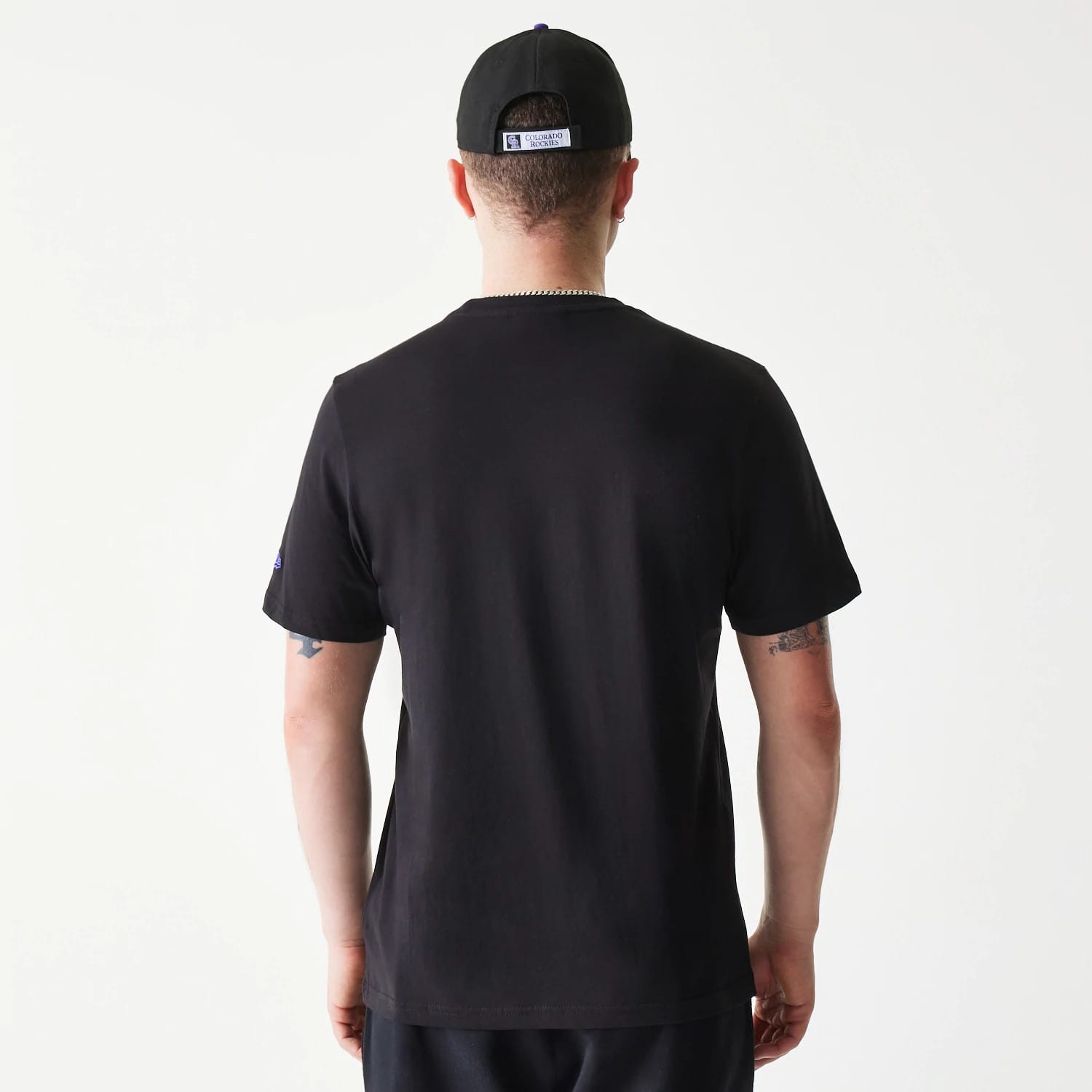 New Era Colorado Rockies MLB Regular Black T-Shirt, on-model rear view showing the solid black back with short sleeves and a raised New Era logo on the sleeve, paired with a black cap featuring a debossed logo on the back strap.