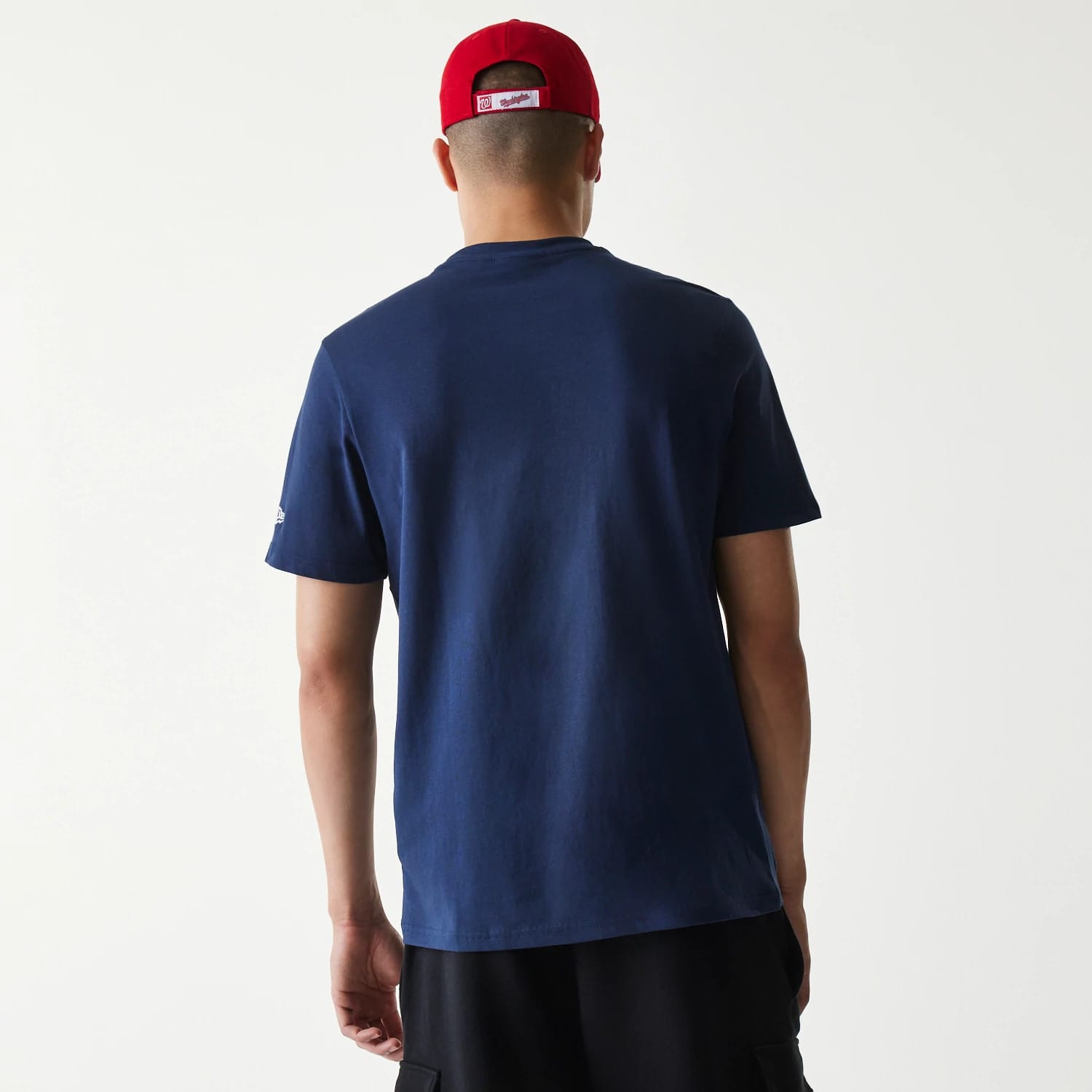 New Era Washington Nationals MLB Regular Dark Blue T-Shirt, rear view showing the plain dark blue fabric of the T-shirt, a debossed New Era logo on the left sleeve, worn by a person in a red cap facing away.