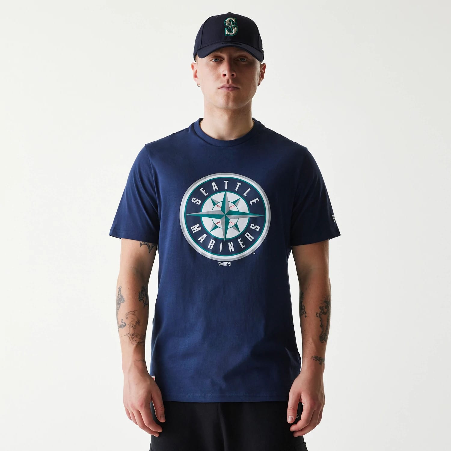New Era Seattle Mariners MLB Regular Dark Blue T-Shirt, on-model view showing a large circular Seattle Mariners logo on the centre of the chest, a navy cap with a raised team emblem, and a small embroidered logo on the left sleeve.