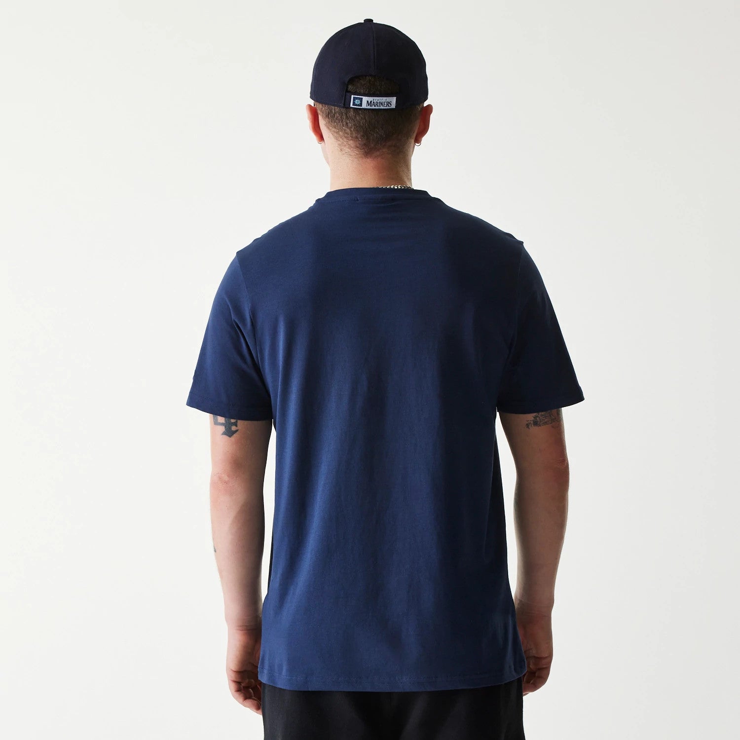 New Era Seattle Mariners MLB Regular Dark Blue T-Shirt, on-model rear view showing the plain dark blue back of the shirt worn by a person, alongside a dark blue cap with a raised Seattle Mariners logo on the rear label.