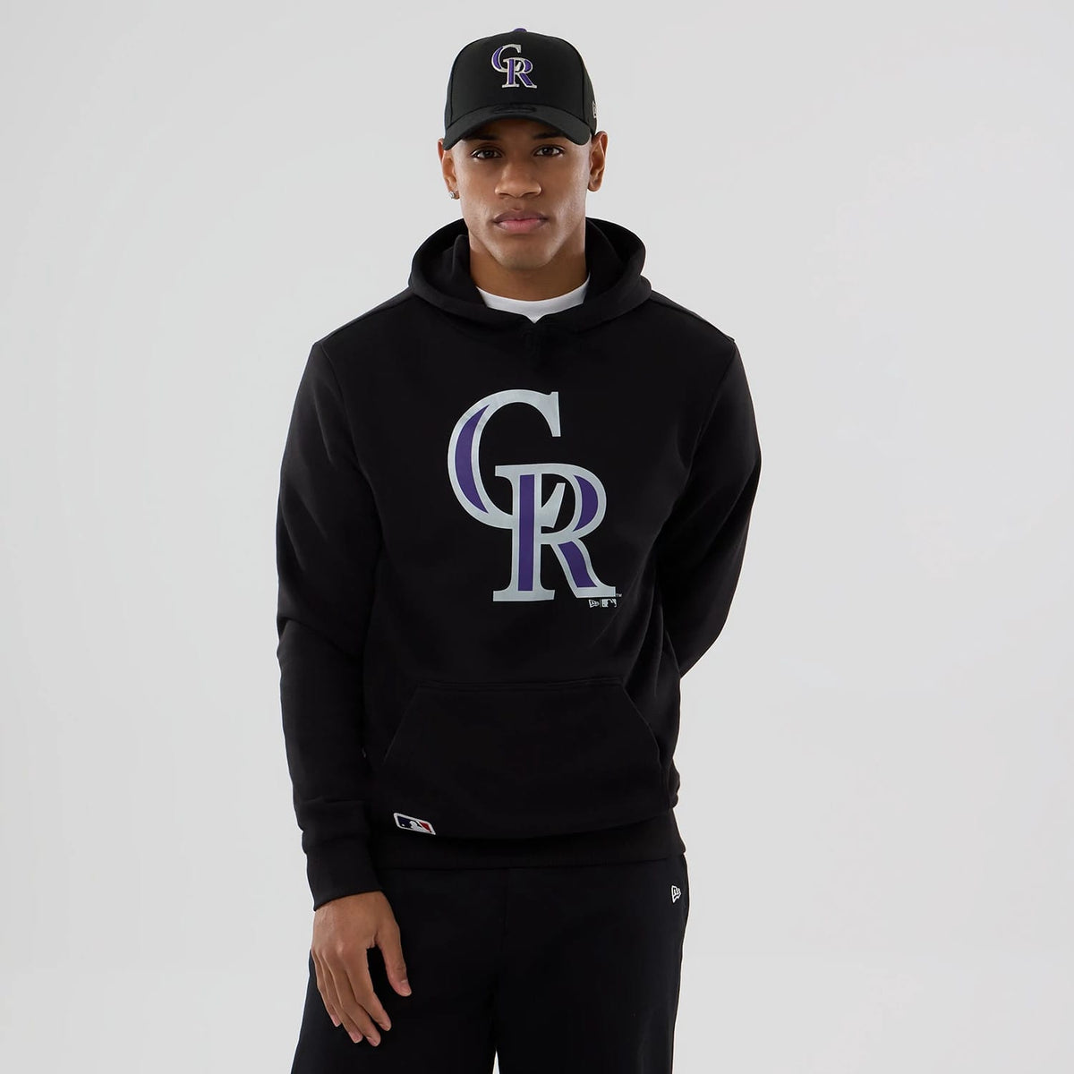 New Era Colorado Rockies MLB Black Pullover Hoodie, front view showing a large 'CR' logo in purple and white on the centre chest, worn by a model also wearing a black cap with a raised 'CR' logo on the front.