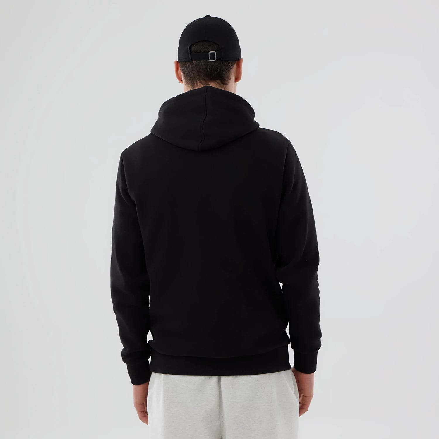 New Era Cincinnati Reds MLB Black Pullover Hoodie, rear view showing the plain black fabric with a large hood, worn by a person also dressed in a black cap and light grey trousers, no logo or additional features visible on the back.