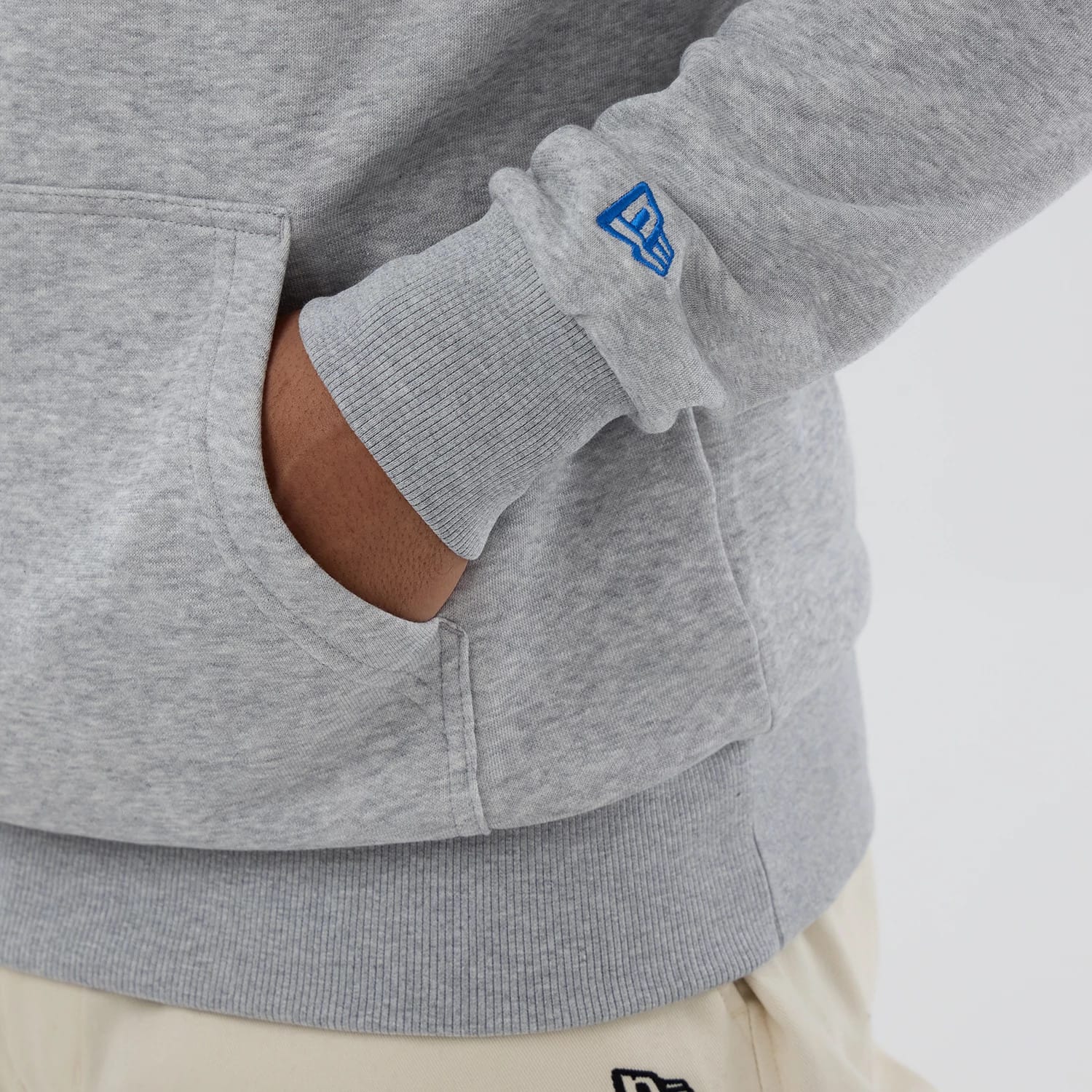 New Era Chicago Cubs MLB Grey Pullover Hoodie, side view showing a ribbed cuff with a blue embroidered New Era logo near the wrist and a grey kangaroo pocket on the front, worn with light beige trousers.