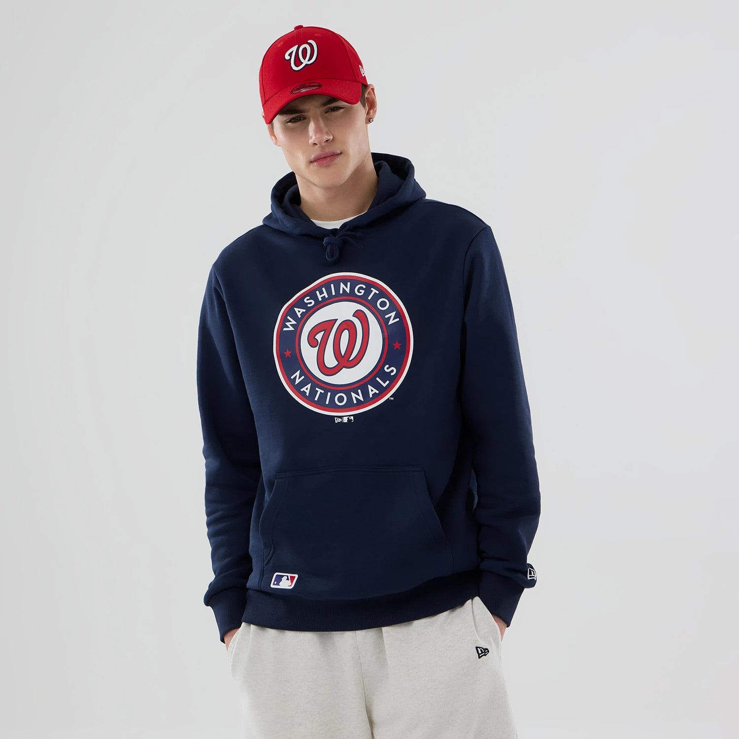 New Era Washington Nationals MLB Dark Blue Pullover Hoodie, on-model view showing the large circular team logo centred on the chest in white and red with a debossed New Era logo on the sleeve, worn with a red cap featuring a raised white team logo.