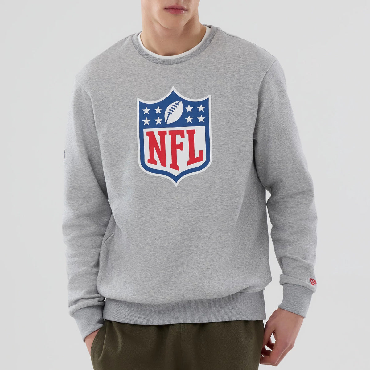 New Era NFL Generic Logo Grey Crew Neck Sweater, front view on a model showing the large red and blue NFL shield logo at the centre of the chest and a small New Era flag logo on the left sleeve cuff.