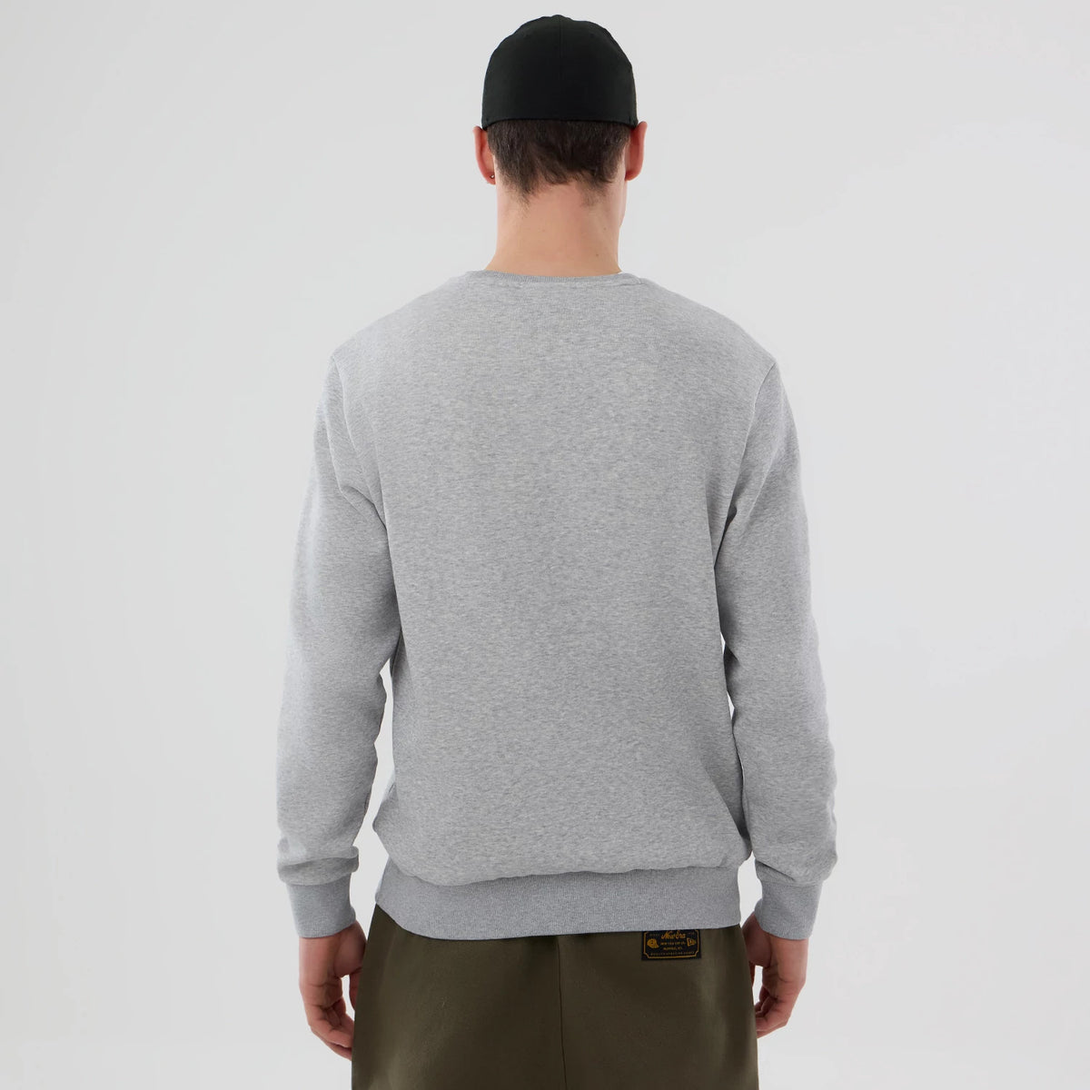 New Era NFL Generic Logo Grey Crew Neck Sweater, back view on a model wearing olive trousers and a black cap, showcasing the simplicity of the light grey fabric and ribbed cuffs, complementing casual style.