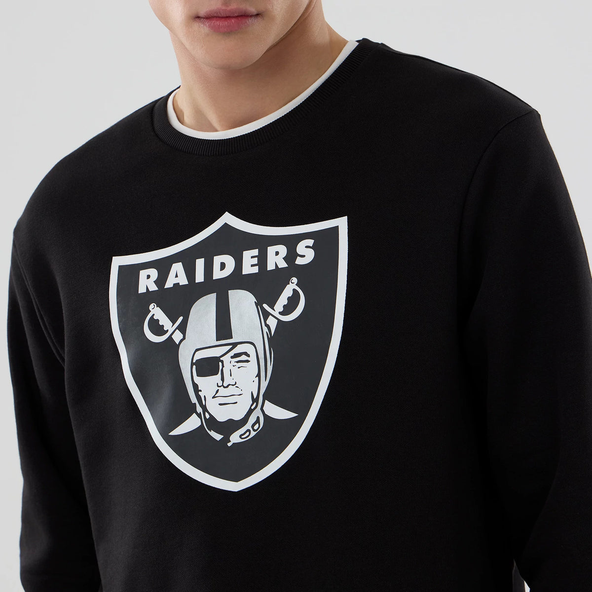 New Era Las Vegas Raiders NFL Core Black Crew Neck Sweater, front view on a model with a large white and grey Raiders logo printed on the chest, featuring ribbed collar detailing with a visible white inner layer.