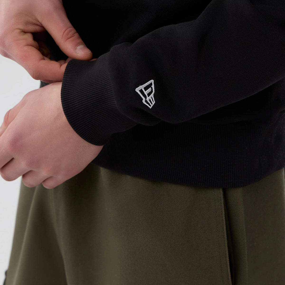 New Era Las Vegas Raiders NFL Core Black Crew Neck Sweater, close-up view with a model showing the white New Era flag logo embroidered on the left sleeve cuff above olive green trousers.