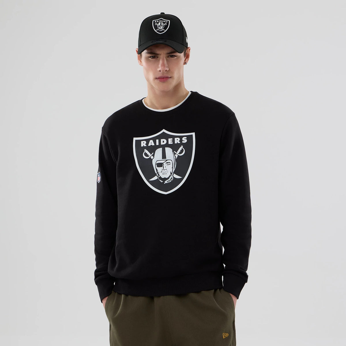 New Era Las Vegas Raiders NFL Core Black Crew Neck Sweater, front view on a model wearing an all-black cap with the Raiders logo, featuring a large white Raiders shield logo on the chest and an NFL patch on the right sleeve.
