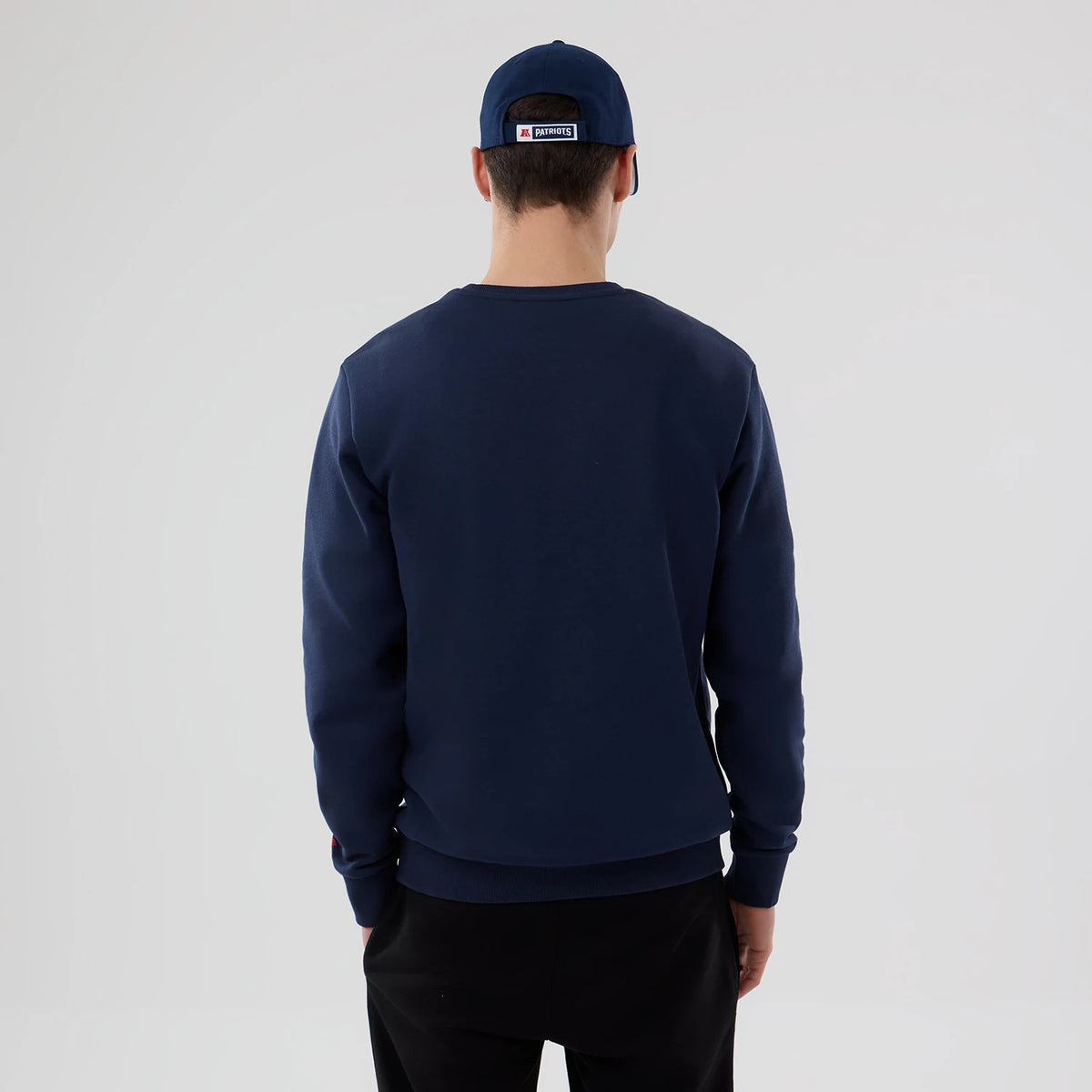 New Era New England Patriots NFL Dark Blue Crew Neck Sweater, back view on a model with a matching dark blue cap, showing the simple solid-colour design and straight hemline across the lower back.
