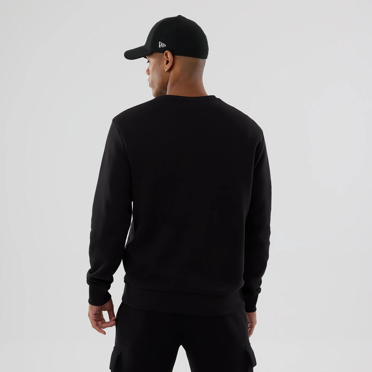 New Era NFL Generic Logo Black Crew Neck Sweater, back view on a model paired with a black cap featuring the New Era flag logo on the left side, showcasing the sweater's plain black back and ribbed cuffs.