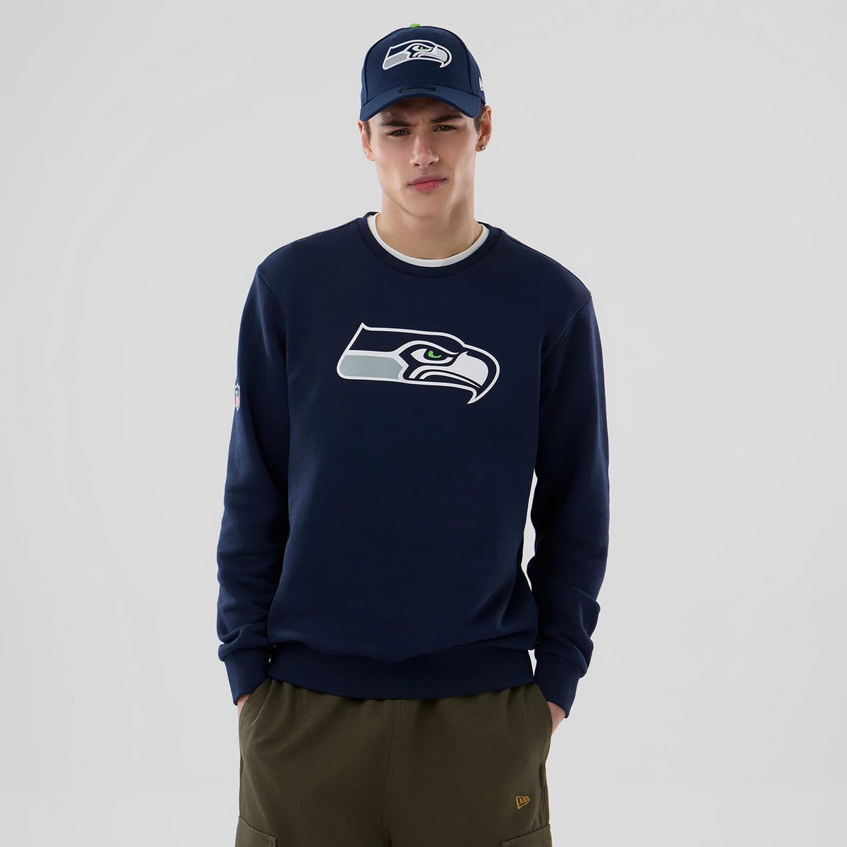 New Era Seattle Seahawks NFL Dark Blue Crew Neck Sweater, front view on a model wearing a matching dark blue cap, displaying the Seahawks logo in white and green centred on the chest, with ribbed cuffs and hem.