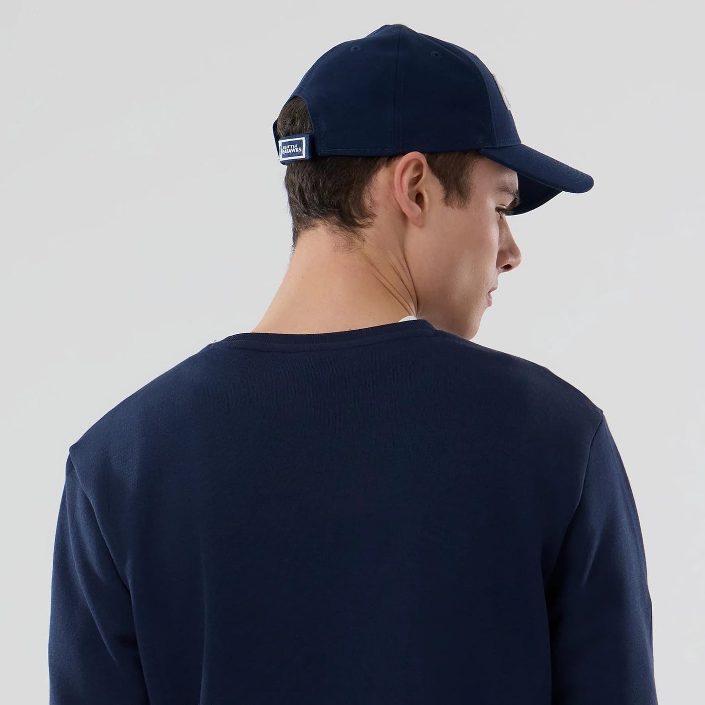 New Era Seattle Seahawks NFL Dark Blue Crew Neck Sweater, back view on a model paired with a dark blue cap, showing a rectangular patch above the opening at the centre back of the cap.