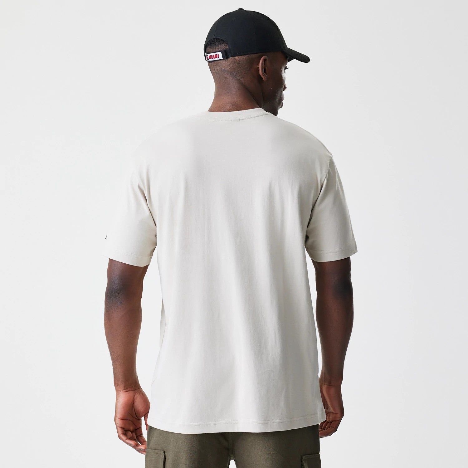 New Era Miami Heat Modern Driver Light Beige T-Shirt, rear view showing the plain light beige fabric with short sleeves, worn by a person also wearing a black cap featuring a small raised Miami logo on the back strap.