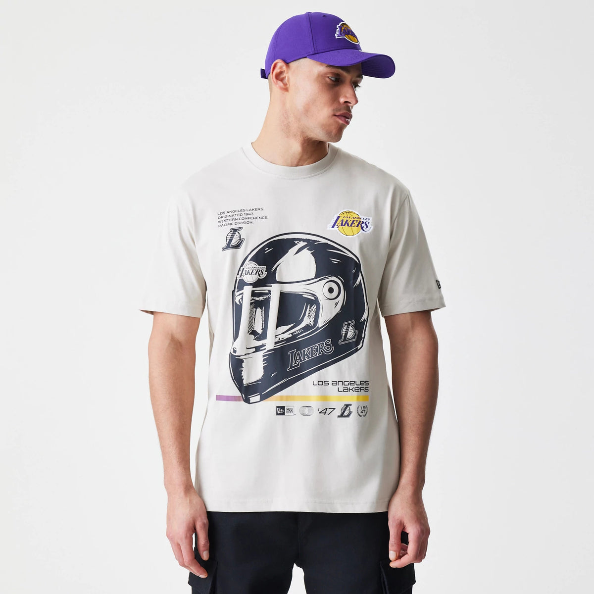New Era LA Lakers Modern Driver Light Beige T-Shirt, front view featuring a large graphic of a helmet with Lakers text and logo in black and white, an embossed Lakers logo on the upper chest, worn by a model in a purple cap with a raised Lakers logo.