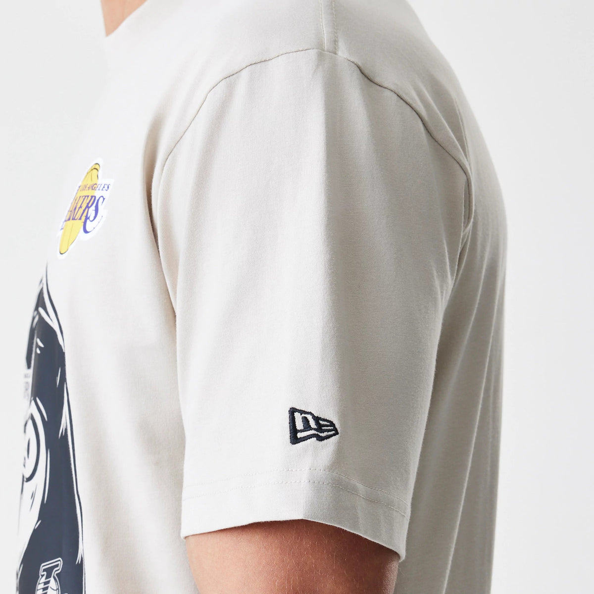 New Era LA Lakers Modern Driver Light Beige T-Shirt, side view showing the light beige short sleeve with a raised New Era logo on the lower arm and part of the printed LA Lakers emblem on the chest.