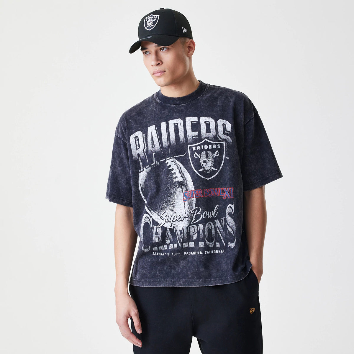 New Era Las Vegas Raiders Sport Classic Black T-Shirt, on-model view showing the front with large Raiders graphics including a football and text, paired with a black cap featuring a raised New Era logo on the side and Raiders emblem on the front.