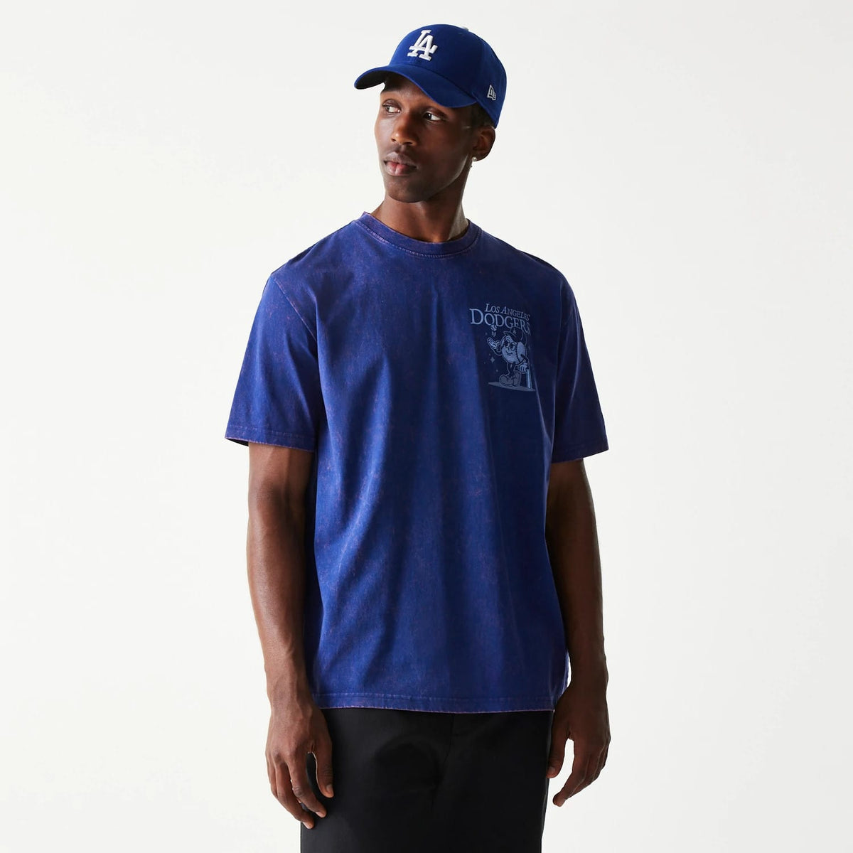 New Era LA Dodgers MLB Character Dark Blue T-Shirt, on-model view showing a dark blue T-shirt with a printed cartoon character and Los Angeles Dodgers text on the chest, paired with a blue cap featuring a raised white LA logo.