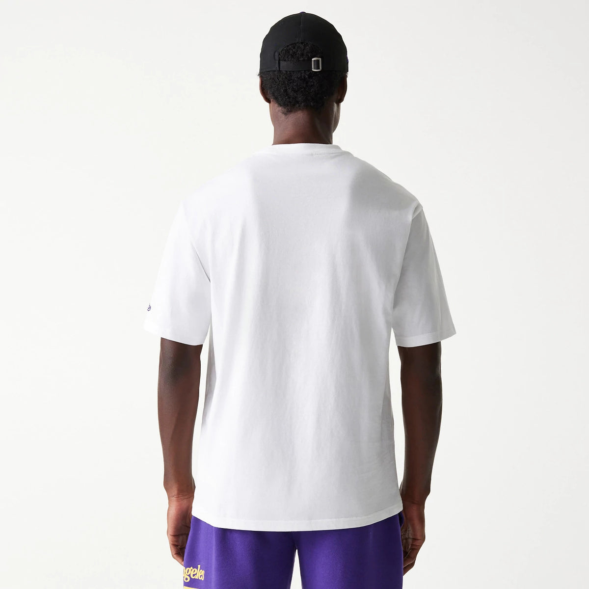 New Era LA Lakers NBA Team Graphic White Oversized T-Shirt, rear view showing a plain white back with short sleeves, a small embroidered logo on the left sleeve and a person wearing a black cap and purple shorts with yellow text.