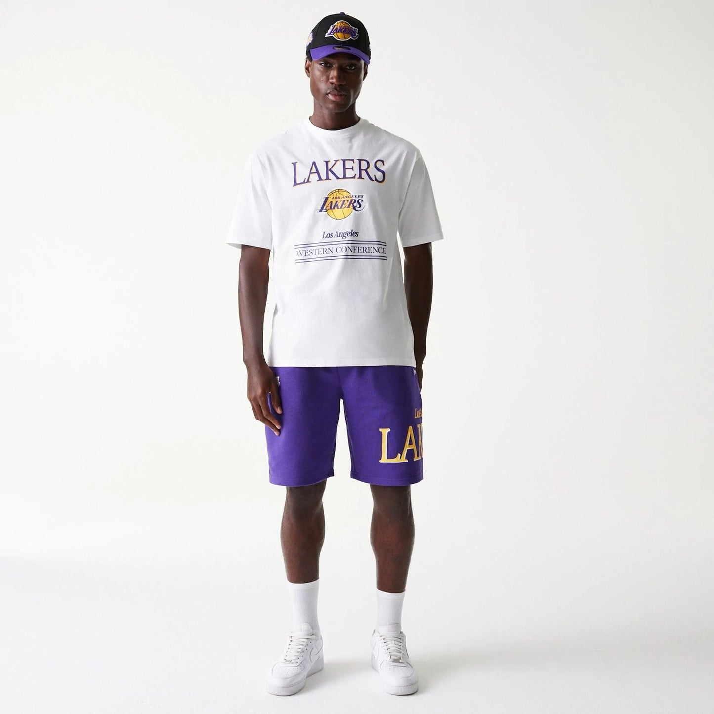 New Era LA Lakers NBA Team Graphic White Oversized T-Shirt, front view showing purple Lakers text and a yellow Los Angeles Lakers logo printed centrally, worn by a model in matching purple shorts and a black cap featuring a raised team logo.