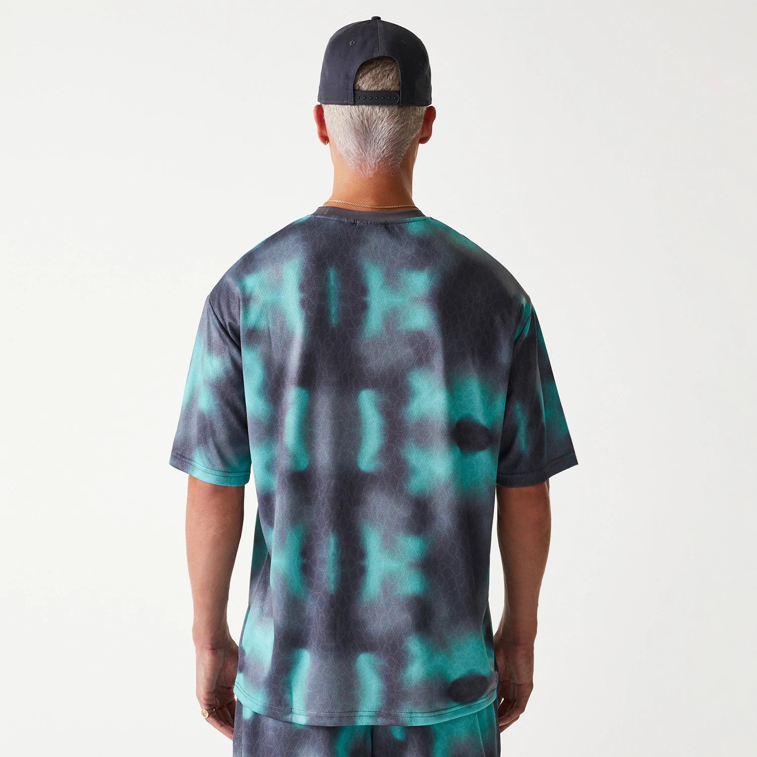 New Era Chicago Bulls NBA All Over Print Green Oversized T-Shirt, rear view showing the green and black tie-dye pattern covering the back, with short sleeves and a relaxed fit, worn by a person with a black cap featuring a row of eyelets on the back.