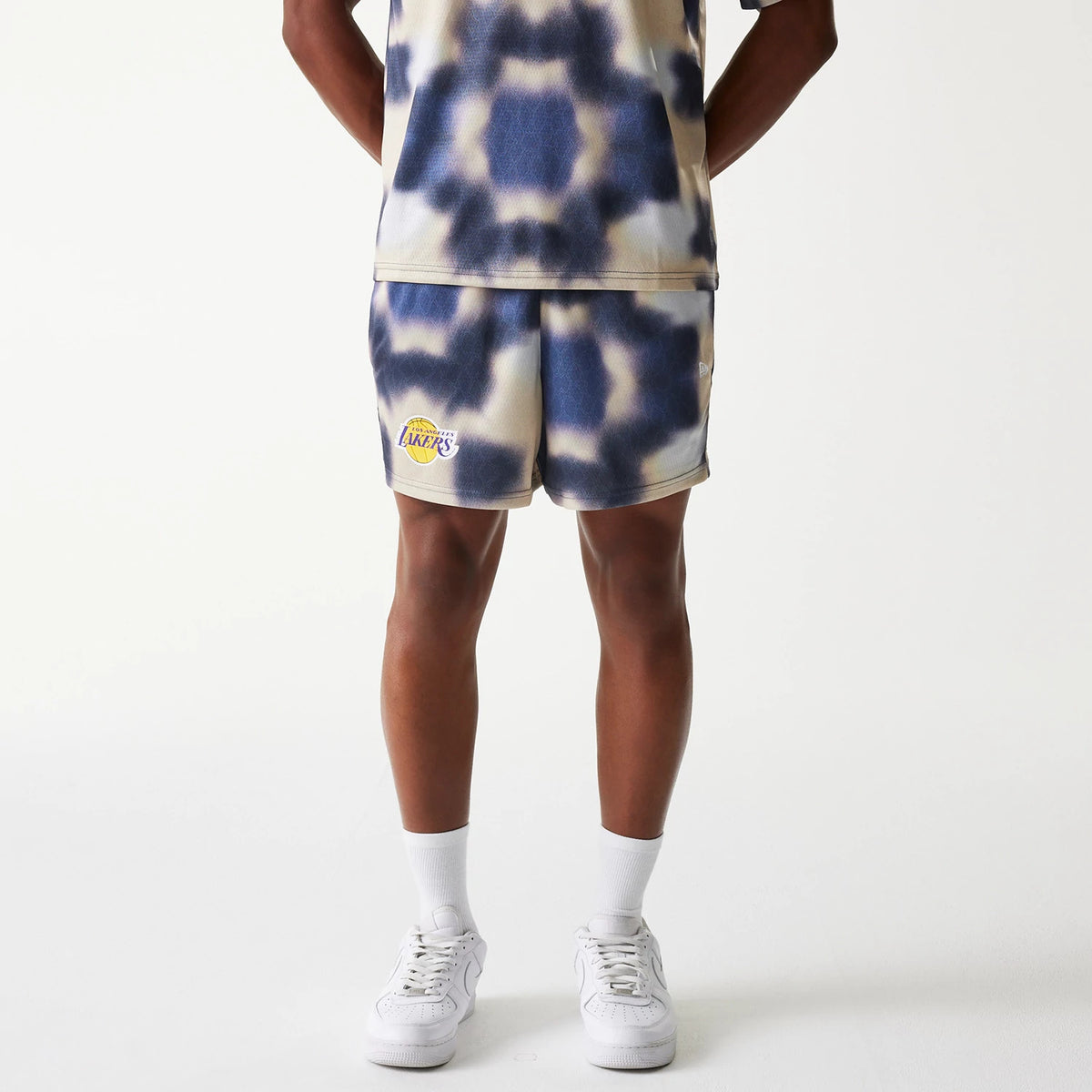 New Era LA Lakers NBA All Over Print Cream Shorts, front view showing the cream and navy tie-dye pattern with a raised Lakers logo on the left leg, paired with matching top, worn by a model standing with feet slightly apart.