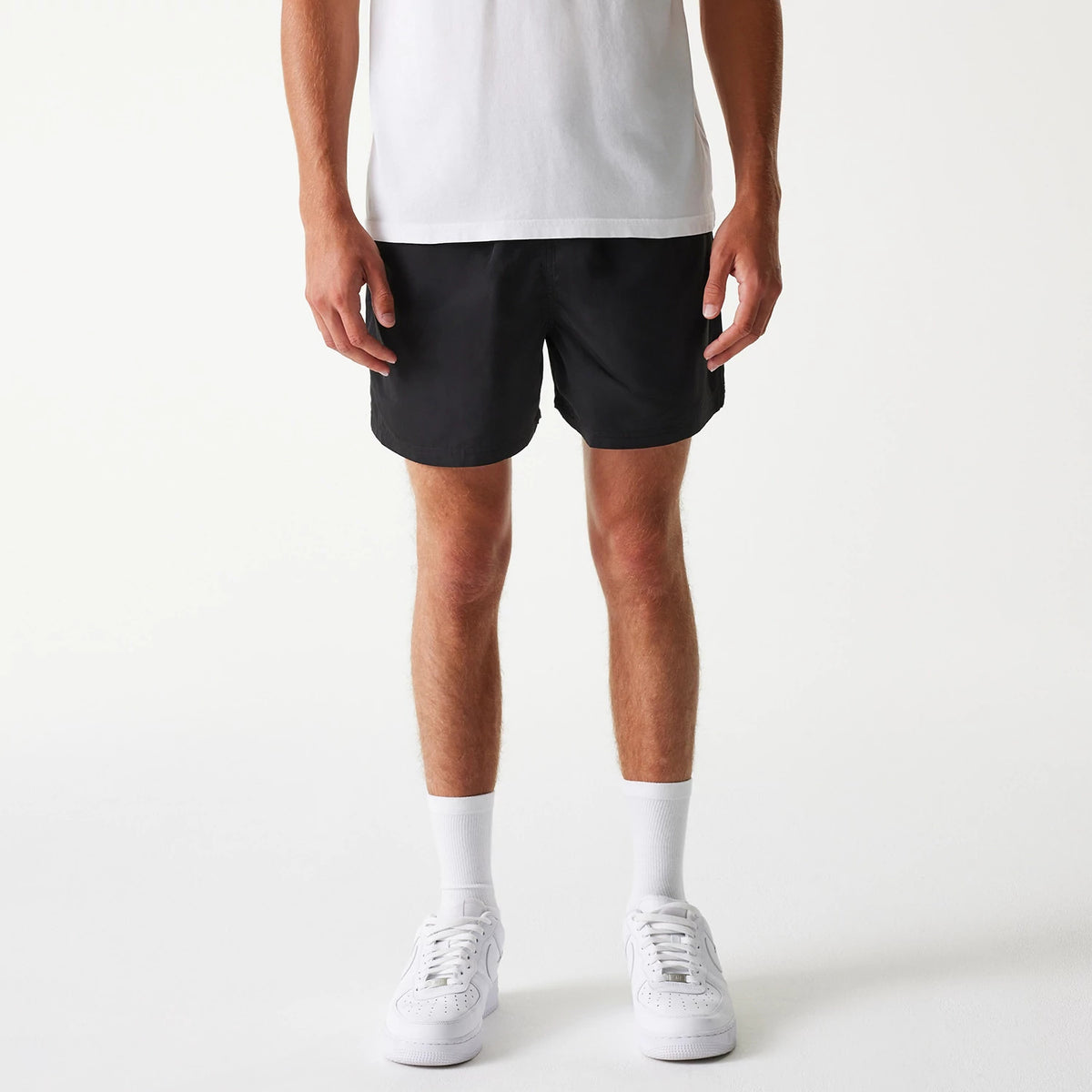 New Era Black Swim Shorts, on-model view showing the plain black shorts with an above-knee length, no visible logos or pockets, paired with a white T-shirt and white trainers.
