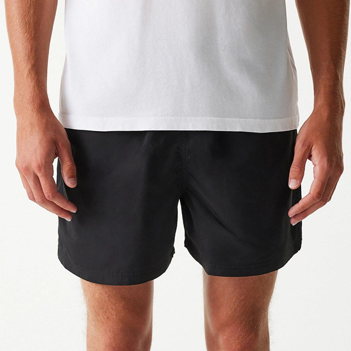 New Era Black Swim Shorts, front view showing the plain black design with a straight hemline and a subtle waistband, worn with a white shirt visible at the top of the image.