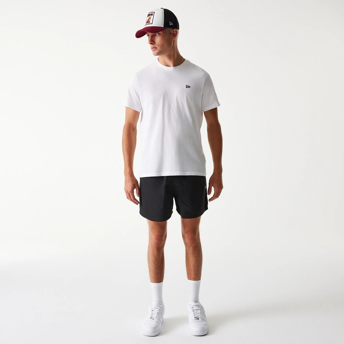 New Era Black Swim Shorts, on-model view showing the black swim shorts with an elasticated waistband paired with a white t-shirt featuring a small raised New Era logo on the chest, and a white and burgundy cap.