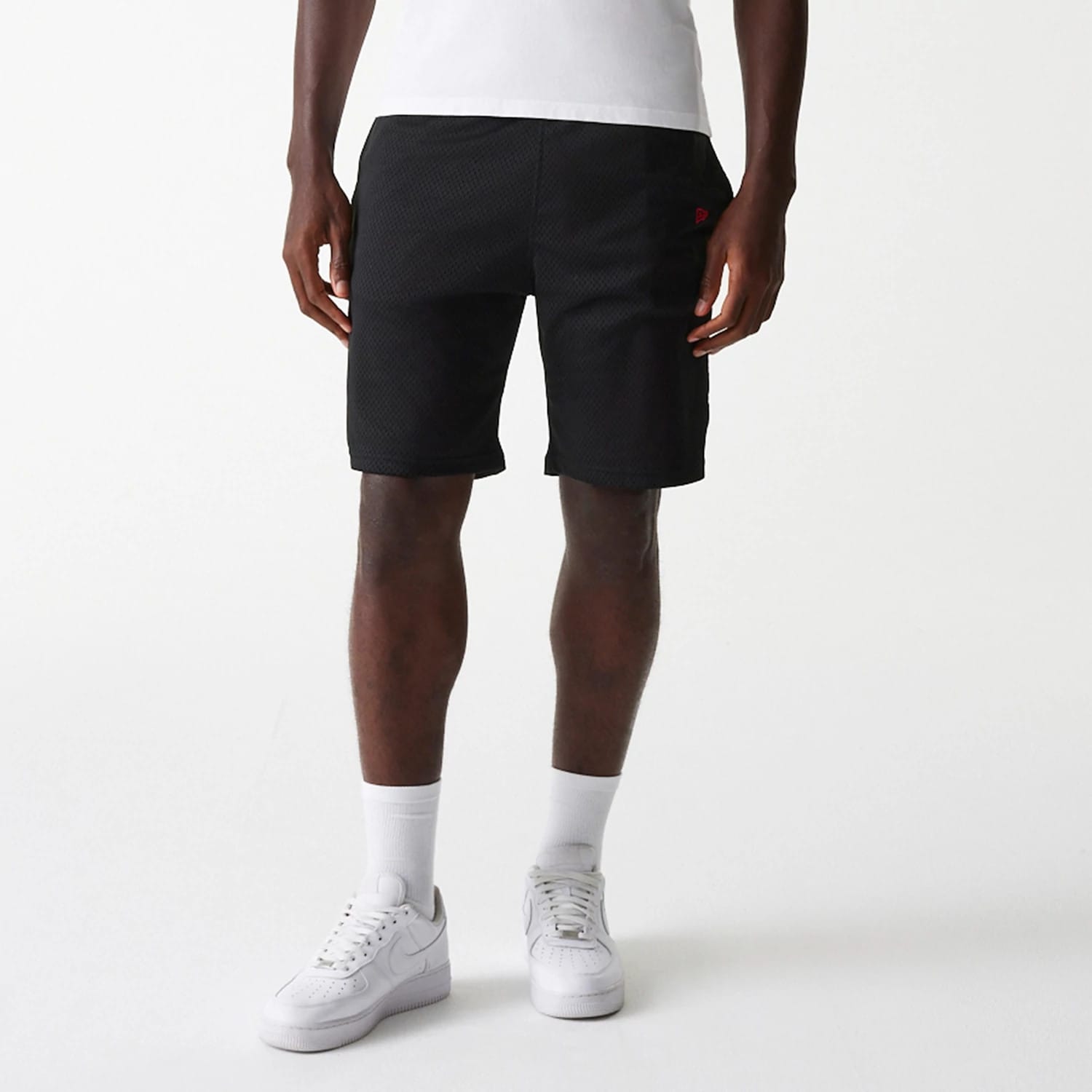 New Era Black Mesh Shorts, front view showing the knee-length black shorts with a raised red New Era logo on the left leg, hands relaxed by the sides, paired with white socks and trainers.