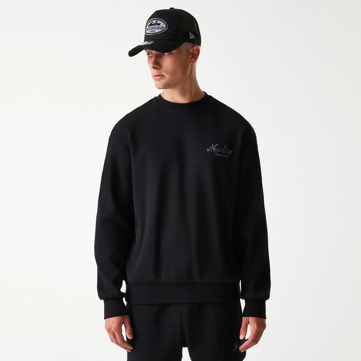 New Era Waffle Black Oversized Crew Neck Sweater, on-model front view showing the embroidered New Era Since 1920 logo on the left chest, paired with a matching black cap featuring a New Era patch and side logo.