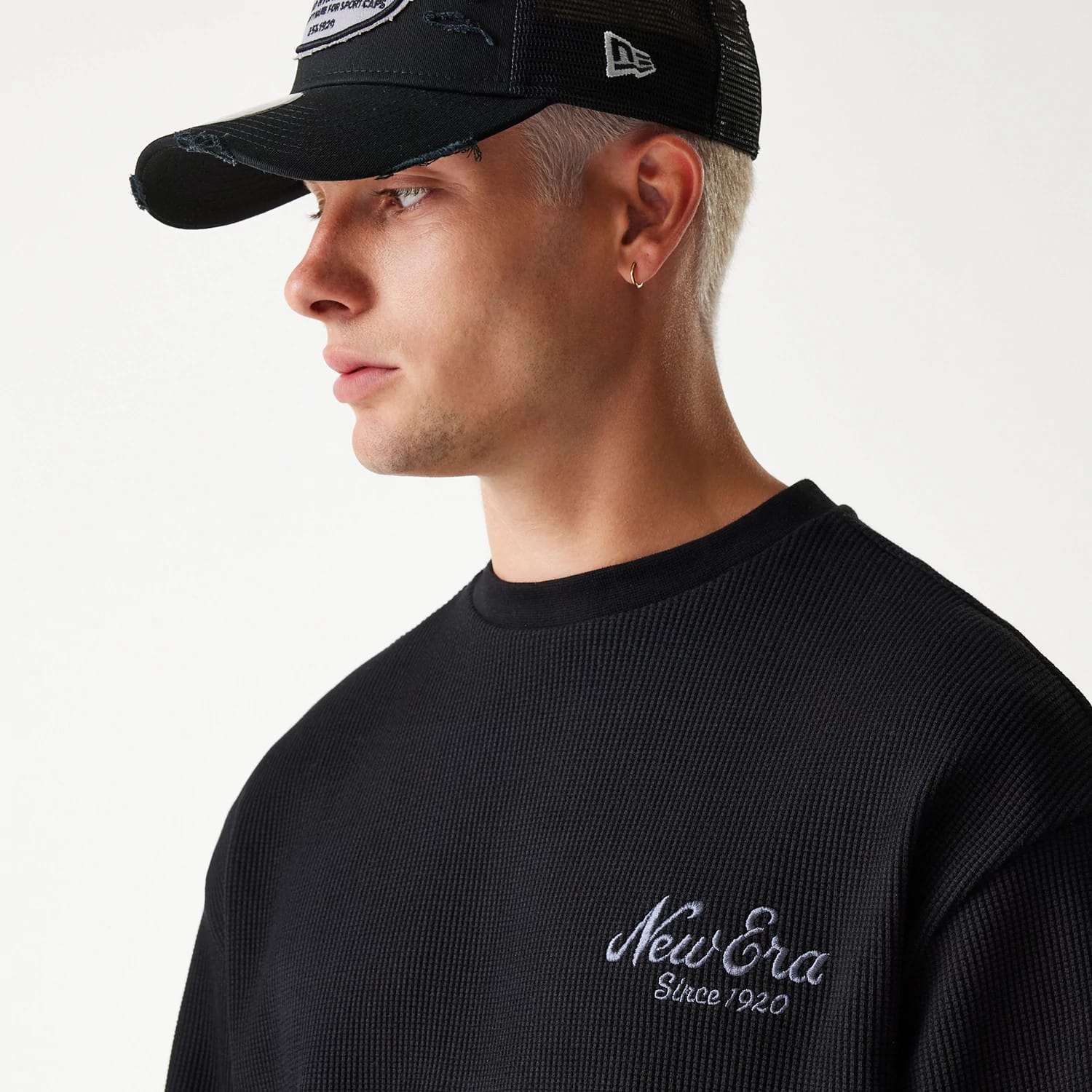 New Era Waffle Black Oversized Crew Neck Sweater, on-model shown with a close-up of the grey embroidered New Era Since 1920 logo on the left chest and textured waffle knit, worn under a black mesh cap with a white embroidered side logo.
