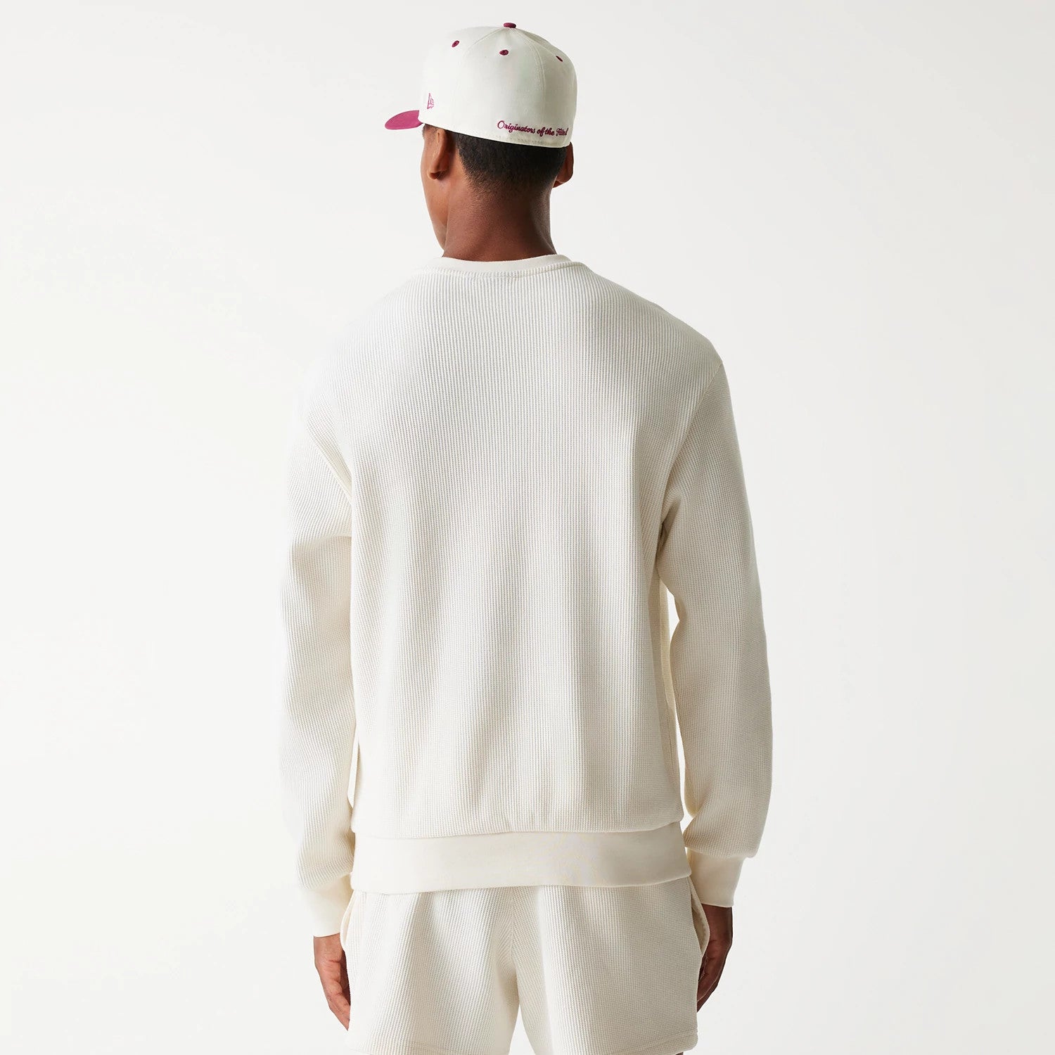 New Era Waffle Open White Oversized Crew Neck Sweater, back view on a model also wearing a white cap with burgundy eyelets, showcasing the ribbed texture and relaxed fit of the sweater over matching shorts.