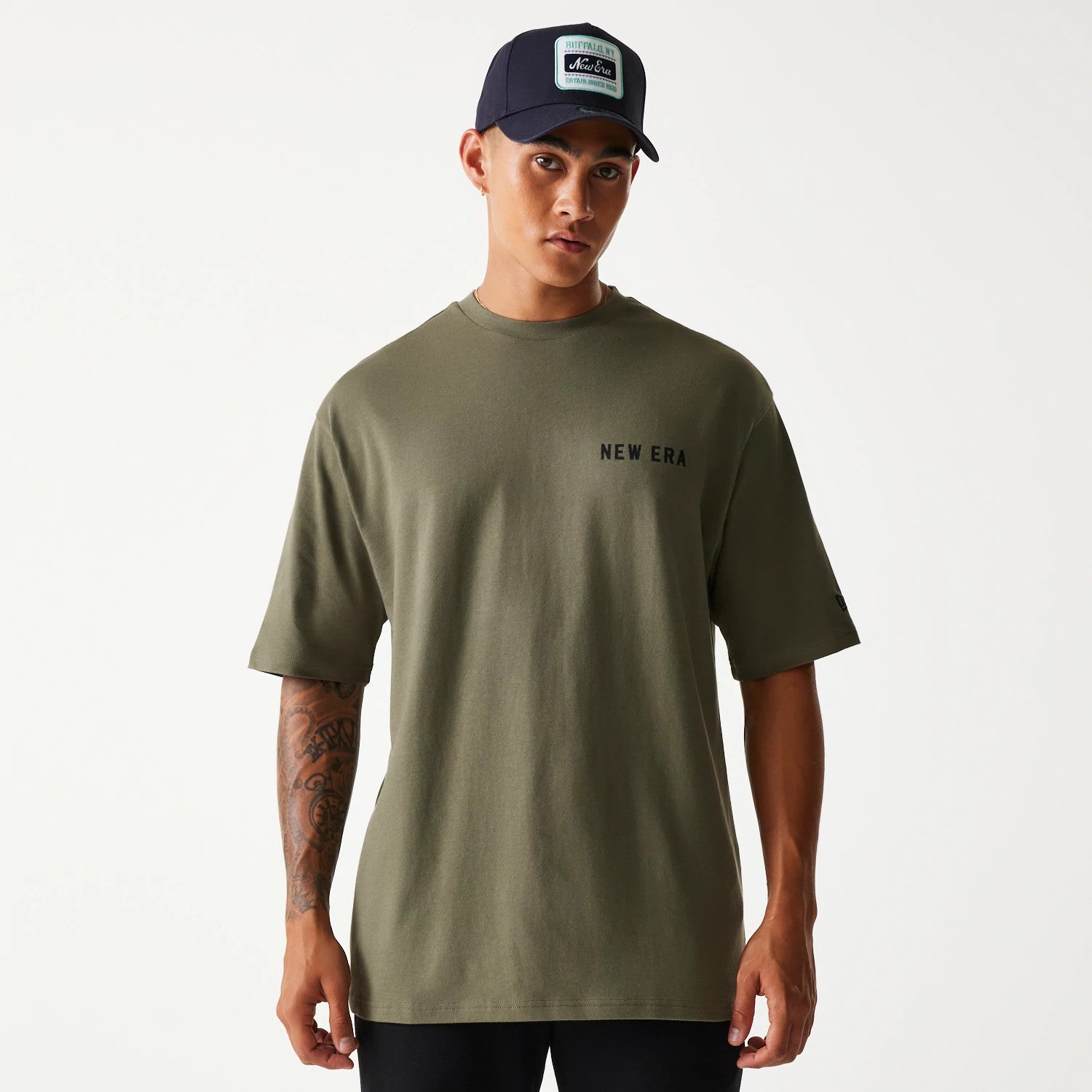 New Era Animal Graphic Green Oversized T-Shirt, on-model view showing the olive green oversized fit with a black NEW ERA text logo on the front left chest, worn with a navy cap featuring a patch logo at the front.