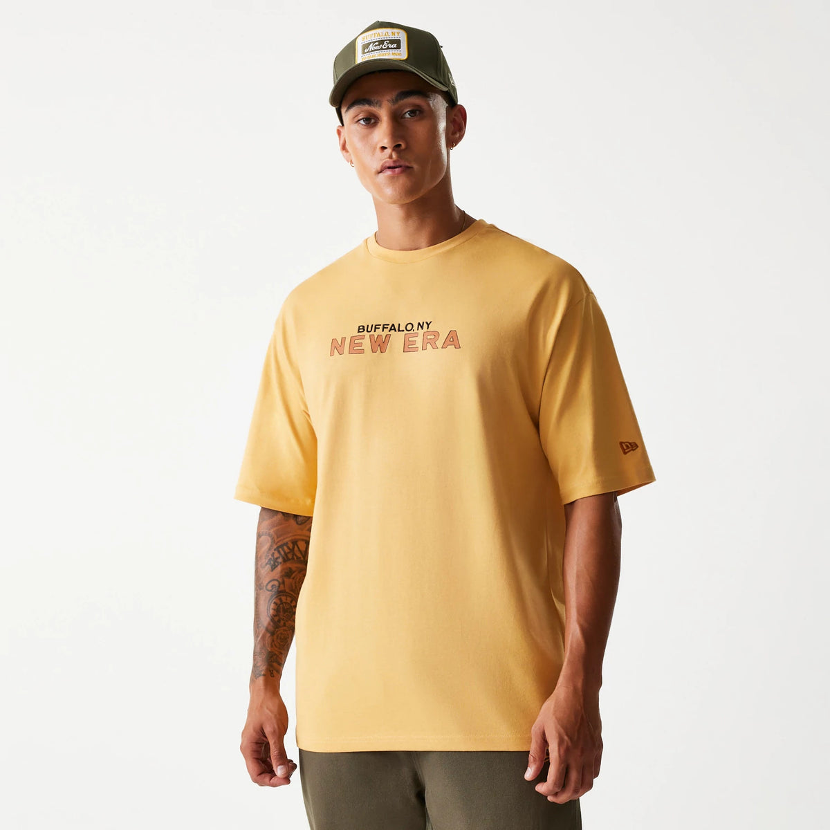 New Era Vacation Graphic Beige Oversized T-Shirt, front view showing the printed Buffalo NY New Era graphic across the chest, a small embroidered New Era logo on the left sleeve, worn by a model with a green cap.