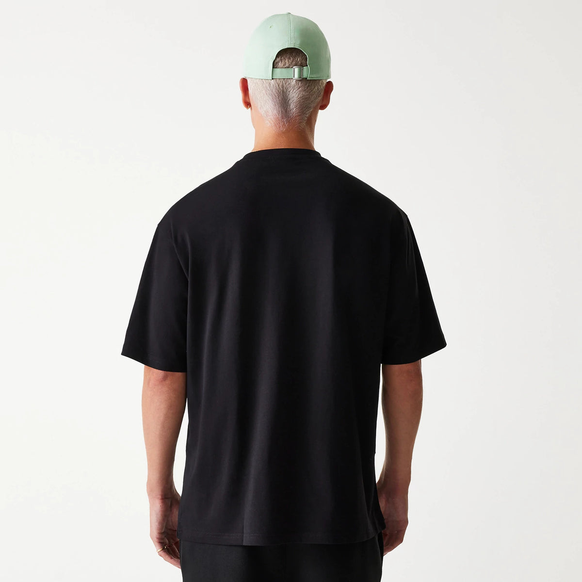 New Era Outdoor Graphic Black Oversized T-Shirt, rear view showing the plain black back panel with short sleeves, worn by a person in a mint green cap with an eyelet and an adjustable strap.