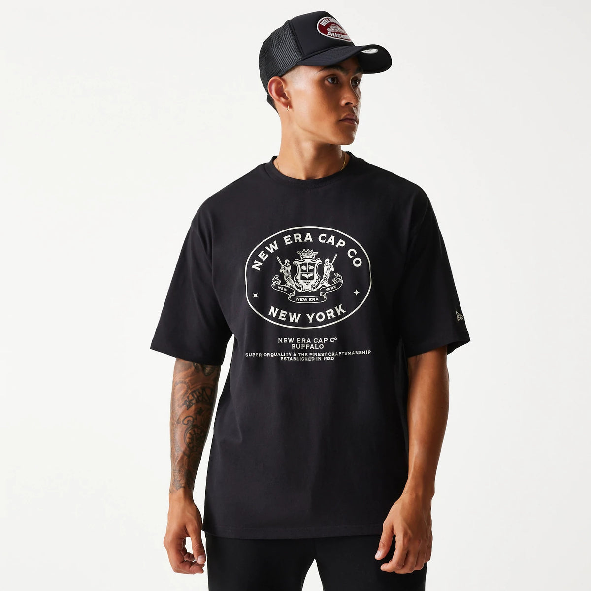 New Era Crest Black Oversized T-Shirt, on-model view showing a large white printed crest logo with text on the centre of the chest, short sleeves, and a small gold debossed New Era flag logo on the left sleeve.
