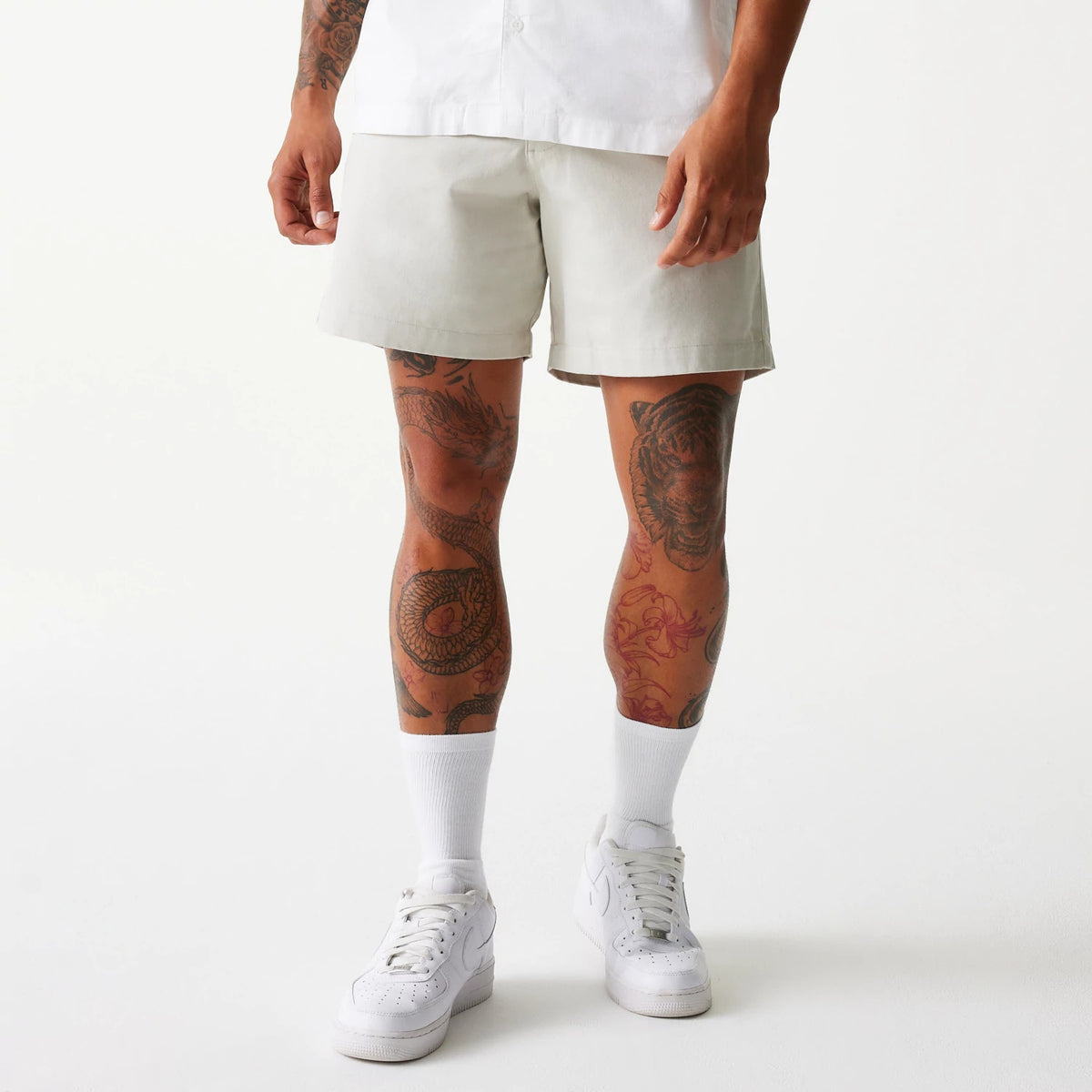 New Era Cream Chino Shorts, on-model view showing the shorts in a light cream colour, mid-thigh length with a straight cut, worn with a white shirt, highlighting tattooed legs and white trainers.