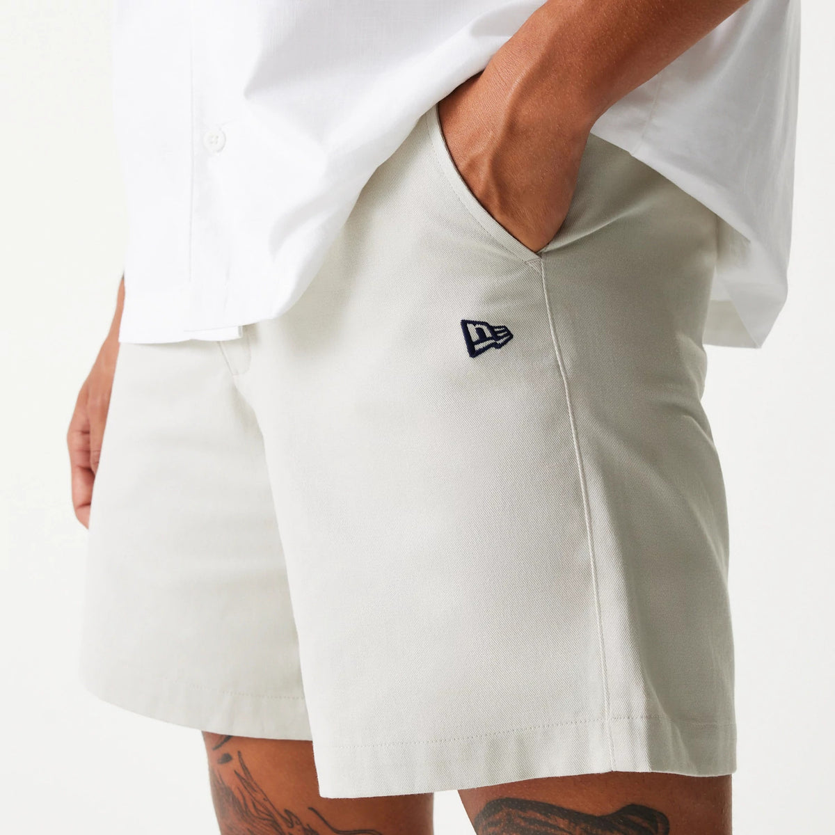 New Era Cream Chino Shorts, side view showing the light cream fabric with a raised New Era logo in dark embroidery near the front pocket, complemented by a crisp white shirt partially tucked in.