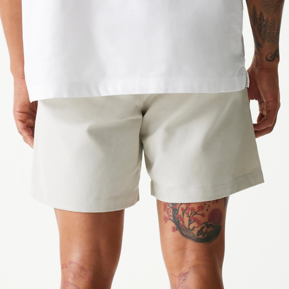 New Era Cream Chino Shorts, rear view showing the straight hemline with a relaxed fit around the thighs, worn by a tattooed model, fabric draping smoothly from the hips to above the knee.